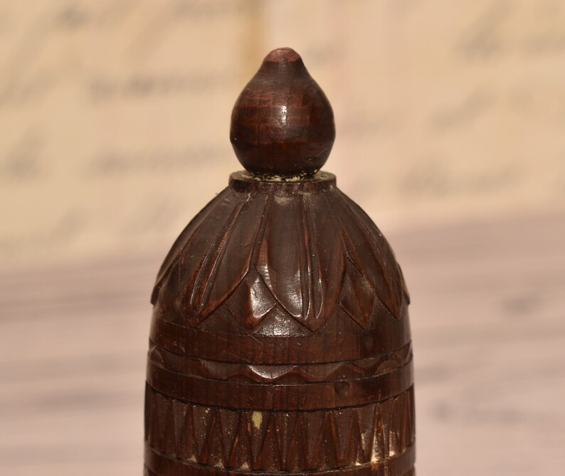 May include: A dark brown wooden finial with intricate carvings. The top of the finial is shaped like a teardrop and the bottom has a series of bands.