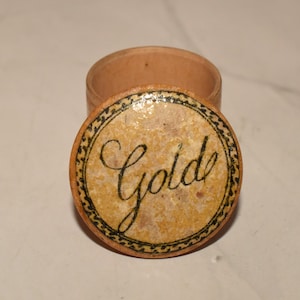 May include: A small, round, open wooden box with a lid. The lid is decorated with the word "Golde" in black script, surrounded by a black and gold border. The box is light brown.