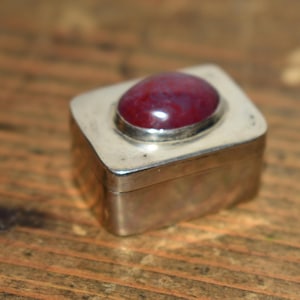May include: A small, square silver box with a large, oval, red gemstone set in the lid. The box is made of silver and has a simple, elegant design. The gemstone is a deep red color.