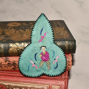 May include: A teal blue fabric bookmark with a black embroidered border. The bookmark is shaped like a leaf and features a small embroidered figure of a person in a pink and yellow outfit holding a small object. The figure is surrounded by embroidered flowers in pink, blue, and green.