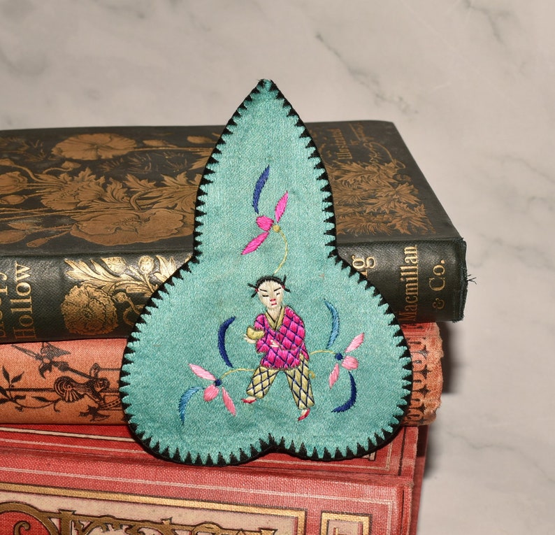 May include: A teal blue fabric bookmark with a black embroidered border. The bookmark is shaped like a leaf and features a small embroidered figure of a person in a pink and yellow outfit holding a small object. The figure is surrounded by embroidered flowers in pink, blue, and green.