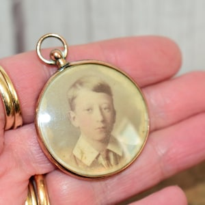 May include: A vintage gold-toned locket with a circular portrait of a young person. The portrait is in sepia tones, and the locket has a small loop at the top for a chain. The locket is held in a hand.
