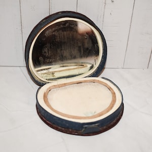 Op de afbeelding: Een open, ovale antieke compact met een spiegel en een poedercompartiment. De compact heeft een donkerblauwe leren buitenkant met een bruine basis. De spiegel is gezet in een witte lijst. Het poedercompartiment heeft een beige stof.