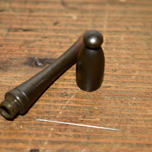 Könnte beinhalten: Ein dunkles Metallwerkzeug-Set auf einer Holzoberfläche. Das Set enthält einen kleinen Hammer, einen zylindrischen Gegenstand und eine dünne, silberne Nadel. Der Hammer hat einen abgerundeten Kopf und einen langen, konischen Griff. Der zylindrische Gegenstand ist dunkelbraun mit einer abgerundeten Oberseite.