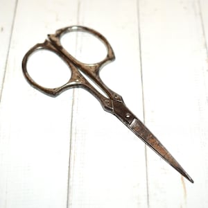 May include: Close-up of antique sewing scissors with a dark, aged metal finish. The scissors have two finger holes and a pointed blade, ideal for detailed cutting tasks. The design features decorative elements near the pivot point.