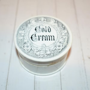 May include: A white, round cold cream container with a black and white floral design. The lid features the words "Cold Cream" in elegant script. The container is set on a white wooden surface.