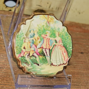 Könnte beinhalten: Ein Vintage-Kompaktspiegel mit goldfarbenem Rahmen und einer gemalten Szene einer Gruppe von Menschen in historischer Kleidung. Die Szene spielt in einem Garten mit Bäumen und einem Zelt. Der Kompaktspiegel befindet sich in einer transparenten Kunststoff-Vitrine.