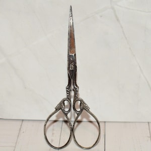 May include: Vintage silver sewing scissors with ornate handles and pointed blades. The scissors have a decorative design on the handles and a slightly rusted blade. The scissors are displayed on a white wooden surface.