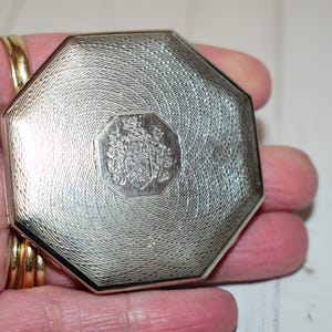 May include: Silver-toned octagonal compact with a textured surface and a central emblem. The compact is held in a hand, showcasing its intricate design and metallic sheen. The emblem appears to be a crest or heraldic symbol.