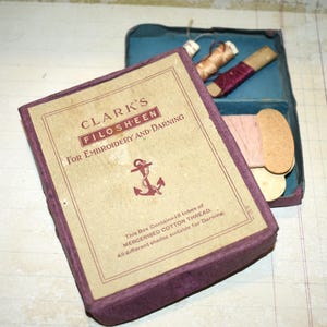 May include: A vintage purple box of Clark's Filosheen thread for embroidery and darning. The box is open, revealing spools of thread in various colors. The box has a label with text and an anchor symbol.