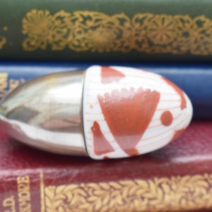 Könnte beinhalten: Ein eiförmiger Behälter mit einem silbernen Metallsockel und einer weiß-roten Musterung. Der Behälter liegt auf einem Stapel Bücher mit verschiedenen Farben und Titeln.