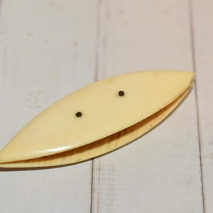 Könnte beinhalten: Eine cremefarbene Vintage-Spule mit zwei kleinen schwarzen Punkten. Die Spule ist oval geformt und scheint aus einem glatten, festen Material zu bestehen. Sie ist etwa 10 cm lang.