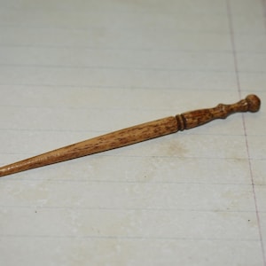Könnte beinhalten: Ein Haarstab aus Holz mit spitzem Ende und dekorativem Griff. Der Stab ist hellbraun mit sichtbarer Holzmaserung. Der Griff hat eine abgerundete Oberseite und geschnitzte Details. Dieses Haaraccessoire dient dazu, das Haar zu einem Dutt zu befestigen.
