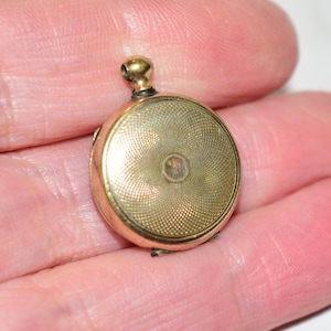 May include: A small, round, gold-toned metal locket featuring a textured, engine-turned guilloché pattern on its surface. The locket has a central circular indentation and a small, rounded bail at the top for attachment to a chain or cord.