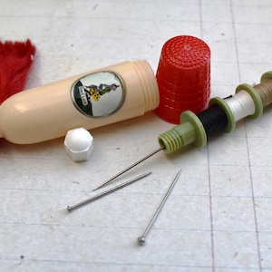 May include: A white plastic sewing kit with a red tassel, a red thimble, and three needles. The kit has a label that says "Just Like" and a picture of a woman in a dress.