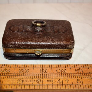 May include: A rectangular, brown leather box with a brass clasp and trim. The box has a textured surface and a brass ring on top. A wooden ruler with centimetre markings is in the foreground.