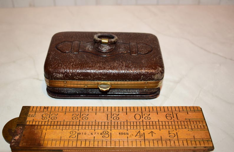 May include: A rectangular, brown leather box with a brass clasp and trim. The box has a textured surface and a brass ring on top. A wooden ruler with centimetre markings is in the foreground.