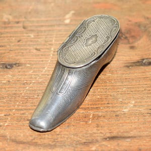 Puede incluir: Una tabaquera de metal plateado con forma de zapato y tapa abatible. La superficie presenta intrincados patrones en relieve, incluyendo un diseño de rejilla texturizada en la tapa superior y detalles lineales a lo largo de la punta y los lados.