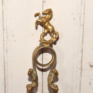 May include: Gold-toned metal fireplace tool with a horse figurine on top. The tool has a ring handle and two curved arms.