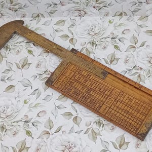May include: A vintage wooden slide rule with a curved end and a rectangular body. The rule has engraved markings and a sliding section with additional calculations. The wood has a warm, aged tone, suggesting its antique nature. The background is a floral pattern.