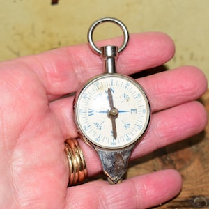 May include: A circular silver-toned metal compass featuring a white face with blue cardinal directions and black degree markings. The device includes a top-mounted metal ring for attachment and a pointed base, displaying a central needle pointer.