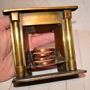 Può includere: Un caminetto in ottone in miniatura con base rettangolare e interni dettagliati. Il caminetto presenta colonne verticali e barre orizzontali, con finitura dorata. Il design è vintage e decorativo.