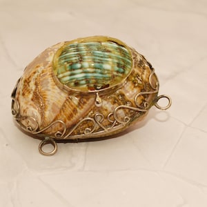 May include: A decorative shell with a silver metal filigree design. The shell features a central oval of green and blue iridescent material. The shell is a natural, organic shape with a silver metal border and small loops.