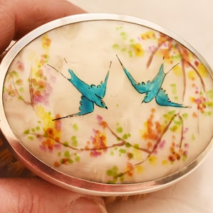 Pode incluir: Escova oval com aro de metal prateado. A escova apresenta um desenho pintado de dois pássaros azuis em voo em meio a ramos florais coloridos. O fundo é de cor creme.