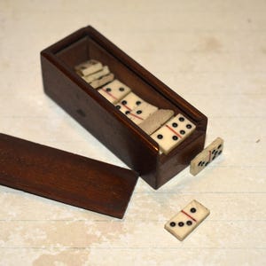 May include: A vintage domino set in a wooden box with a lid. The dominoes are cream-coloured with black dots and a red line. The box is dark brown and rectangular, with the lid open beside it.