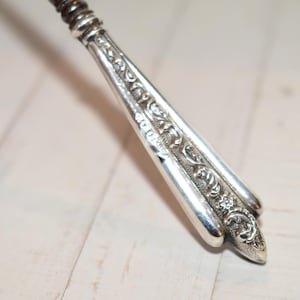 Puede incluir: Un mango de utensilio de metal plateado con un intrincado patrón floral y de volutas en relieve a lo largo de su longitud. El mango se estrecha hacia un extremo puntiagudo y muestra una serie de pequeños sellos grabados en el borde.