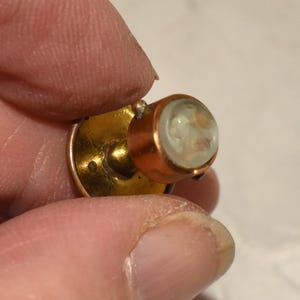 May include: A vintage gold-coloured button with a copper-coloured bezel. The button features a small, round, clear glass or crystal cabochon with an abstract design. The button is held between fingers.