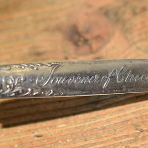 May include: A silver-toned metal souvenir spoon handle featuring an engraved floral border and the cursive text Souvenir of Chicago. The handle has a polished finish with decorative scrollwork detailing along the edges and a rounded end.