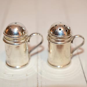 May include: Two vintage silver salt and pepper shakers. Each shaker has a small handle and a domed lid. One lid has a single hole, the other has multiple holes. The shakers are on a white wooden surface.