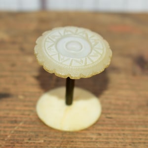 Peut inclure: Un bouchon de bouteille décoratif blanc cassé avec un motif circulaire détaillé. Il possède une tige sombre et une base ronde. Le motif comprend un cercle central et des motifs géométriques. L'objet est posé sur un fond en bois.