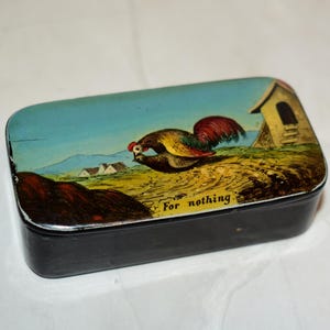 May include: A rectangular black tin box with a colourful lid featuring a rooster illustration. The lid depicts a rooster, a small building, and the text "For nothing" against a blue sky and yellow landscape.
