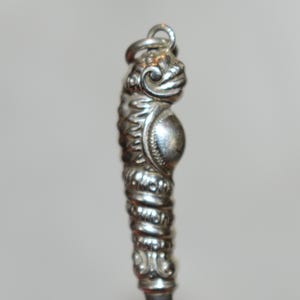 May include: A silver-toned metal charm or spoon handle featuring a sculpted creature with a rounded belly, textured scales, and a prominent head. The object is topped with a small circular jump ring for attachment, showcasing intricate relief detailing throughout the metallic surface.