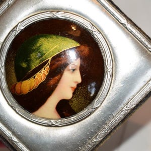 May include: A vintage, silver-toned compact mirror with a central, circular portrait of a woman in profile. The portrait features a woman with auburn hair and a green hat with gold accents. The compact has a square shape with a decorative border.