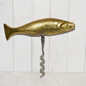 May include: A gold-toned metal corkscrew featuring a handle shaped like a fish with a textured scale pattern. The corkscrew mechanism is made of polished silver-toned metal and extends downward from the center of the fish-shaped handle.