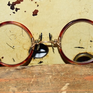 May include: A pair of vintage pince-nez spectacles featuring round, tortoiseshell-patterned rims in shades of brown and amber. The frames are connected by a thin, metallic gold-toned bridge with adjustable nose pads and small metal hardware.