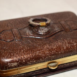 May include: A rectangular brown leather case with a textured surface and a gold-coloured metal frame. The case features an embossed design on the top and a ring-shaped handle. The clasp is also gold-coloured.
