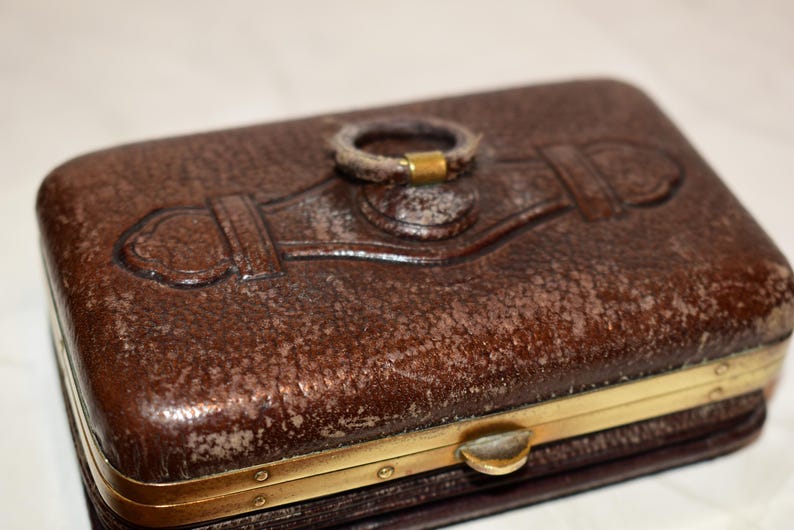 May include: A rectangular brown leather case with a textured surface and a gold-coloured metal frame. The case features an embossed design on the top and a ring-shaped handle. The clasp is also gold-coloured.