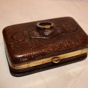 May include: A rectangular brown leather box with a textured surface and gold-coloured metal trim. The box features a decorative ring on the top and a clasp closure. The box is likely used for storing small items.
