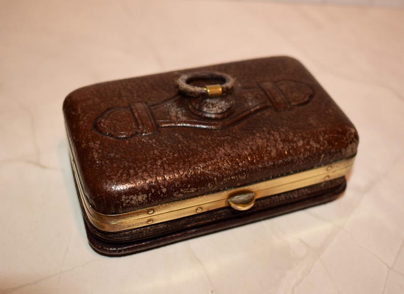 May include: A rectangular brown leather box with a textured surface and gold-coloured metal trim. The box features a decorative ring on the top and a clasp closure. The box is likely used for storing small items.