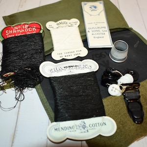May include: A collection of sewing supplies, including spools of black thread, buttons, a thimble, and a needle case. The thread spools have labels with text, including "SHAMROCK" and "MENDING COTTON". The items are arranged on a dark green and black fabric.