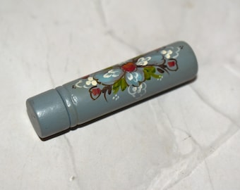 May include: A cylindrical, hand-painted wooden object in a muted blue-gray hue. The object features a floral design with light blue, red, and green accents. The painted flowers and leaves are arranged around the cylinder.