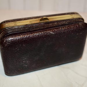 May include: A vintage, rectangular, dark brown leather wallet with a textured surface. The wallet has a gold-coloured metal frame and clasp. The wallet is slightly open, revealing the interior.