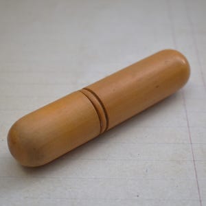 May include: A light brown, wooden needle case. The cylindrical case has rounded ends and a groove near the center. The wood grain is visible, and the case appears to be made of a single piece of wood.