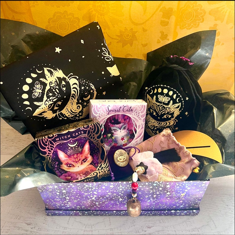 Pode incluir: Uma caixa de presente roxa com um padr&atilde;o de estrelas cheia de v&aacute;rios itens com tema de gato, incluindo uma bolsa de veludo preto com um design de lua e estrelas douradas, um cart&atilde;o roxo e dourado com o texto "Witch Cats", um cart&atilde;o roxo e dourado com o texto "Spirit Cats", e uma bolsa roxa e dourada com um tassel.