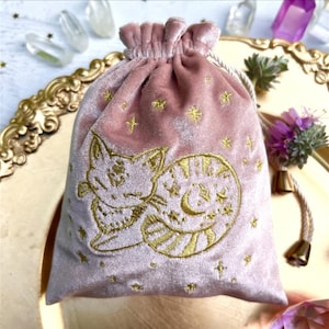 Puede incluir: Una bolsa de terciopelo rosa con cordón y bordado dorado de un diseño de gato celestial. La bolsa presenta una parte superior fruncida y un cierre de cordón dorado. Estrellas doradas están dispersas por la superficie de la bolsa. La bolsa probablemente se usa para guardar objetos pequeños.