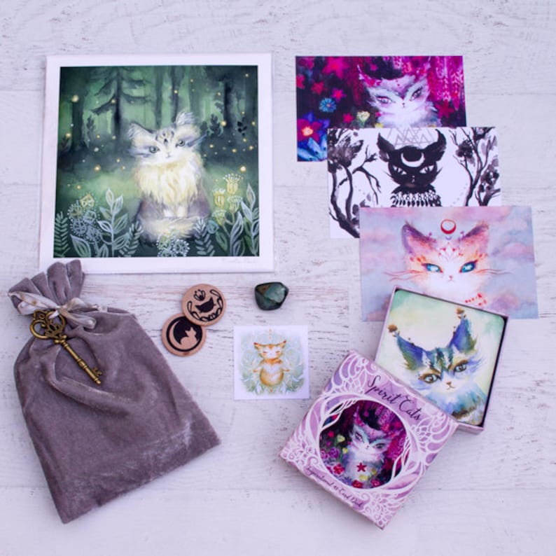 Pode incluir: Um conjunto de cartas de or&aacute;culo Spirit Cats em uma caixa roxa com um gato branco na frente. A caixa &eacute; cercada por v&aacute;rios outros itens, incluindo uma pequena pintura de um gato em uma floresta, uma pequena bolsa com uma chave e alguns cart&otilde;es pequenos com ilustra&ccedil;&otilde;es de gatos.